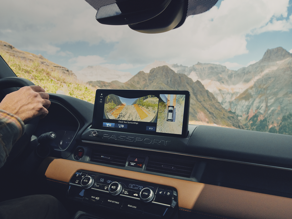 The hands of a person driving a Honda Passport are visible on the steering wheel. To the right, a touchscreen display shows a video feed backup camera. The dashboard is finished with a light brown leather and contains dials, buttons, and air vents.